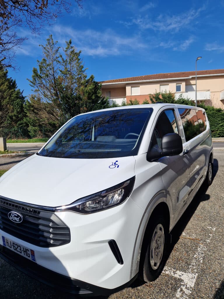 Ford Transit TPMR : Van Blanc avec Logo Accessibilité Van Ford blanc aménagé TPMR avec logo handicapé sur le capot, stationné sous un ciel bleu ensoleillé.