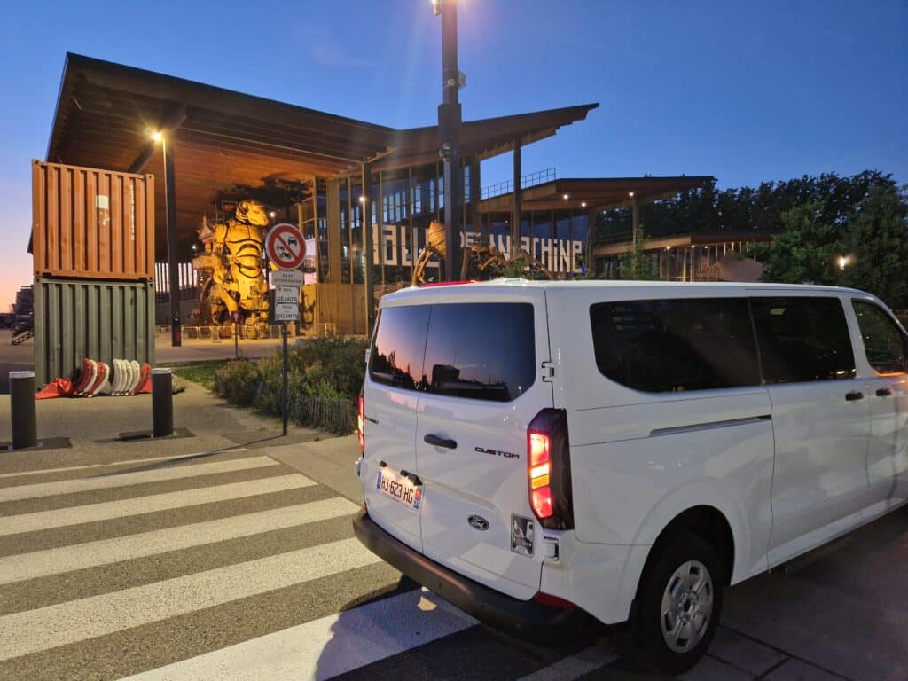 Van blanc Ford Custom devant la Halle de la Machine à Toulouse, avec le Minotaure géant en arrière-plan au crépuscule.