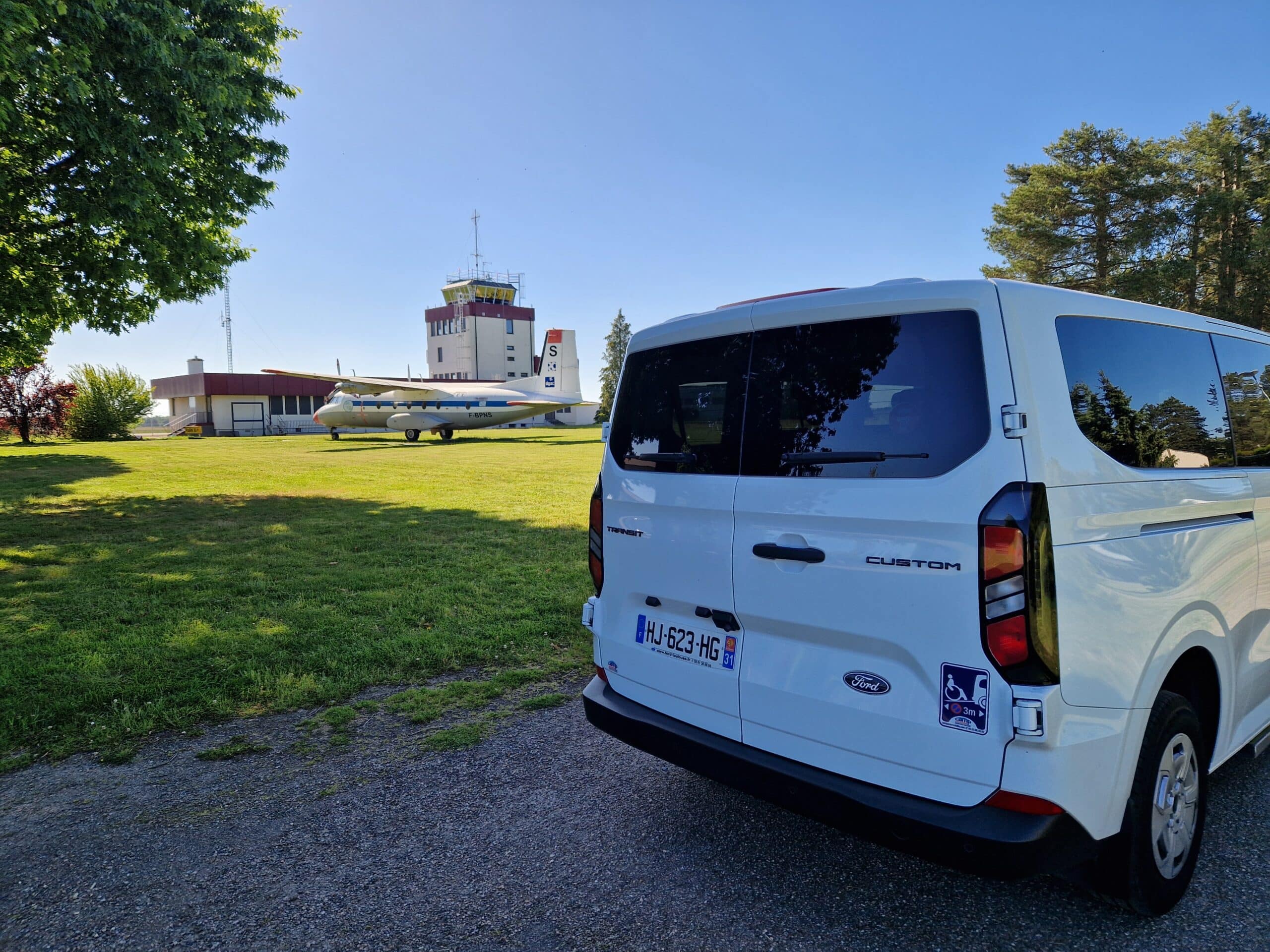 Ford Transit Custom blanc à l’aérodrome avec avion Ford Transit Custom blanc HJ-623-HG garé près d'un avion F-BPNS et d'une tour de contrôle par temps clair.