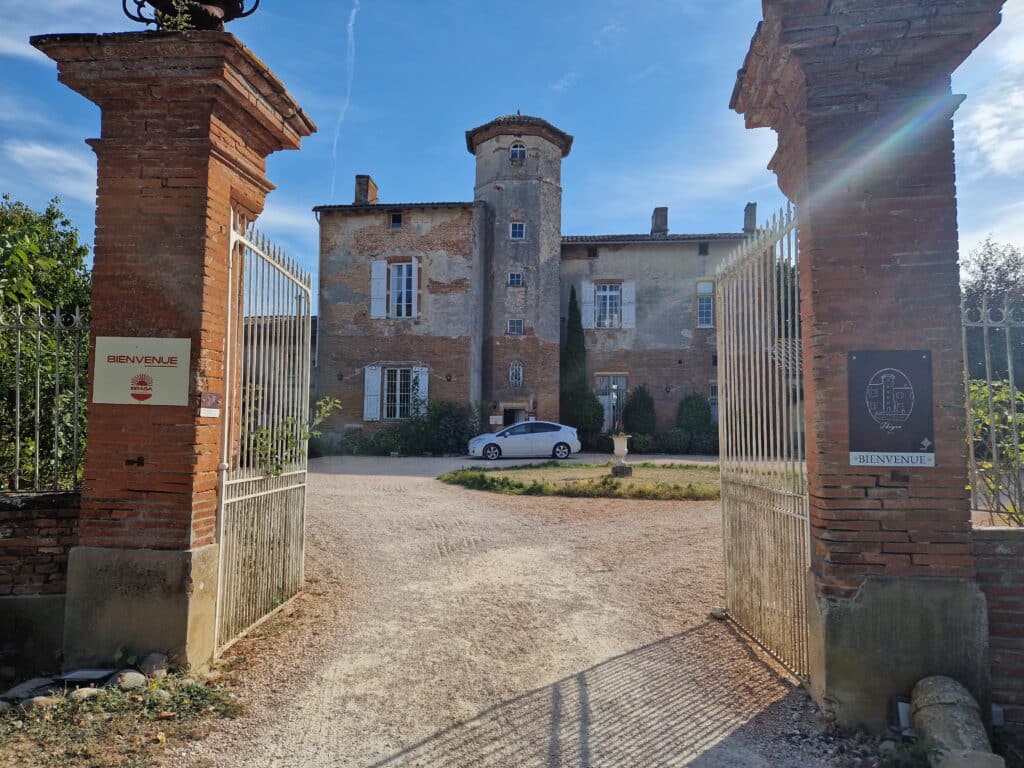Entrée du Château de Thégra en briques rouges, portail ouvert et panneaux Bienvenue sous un ciel bleu.