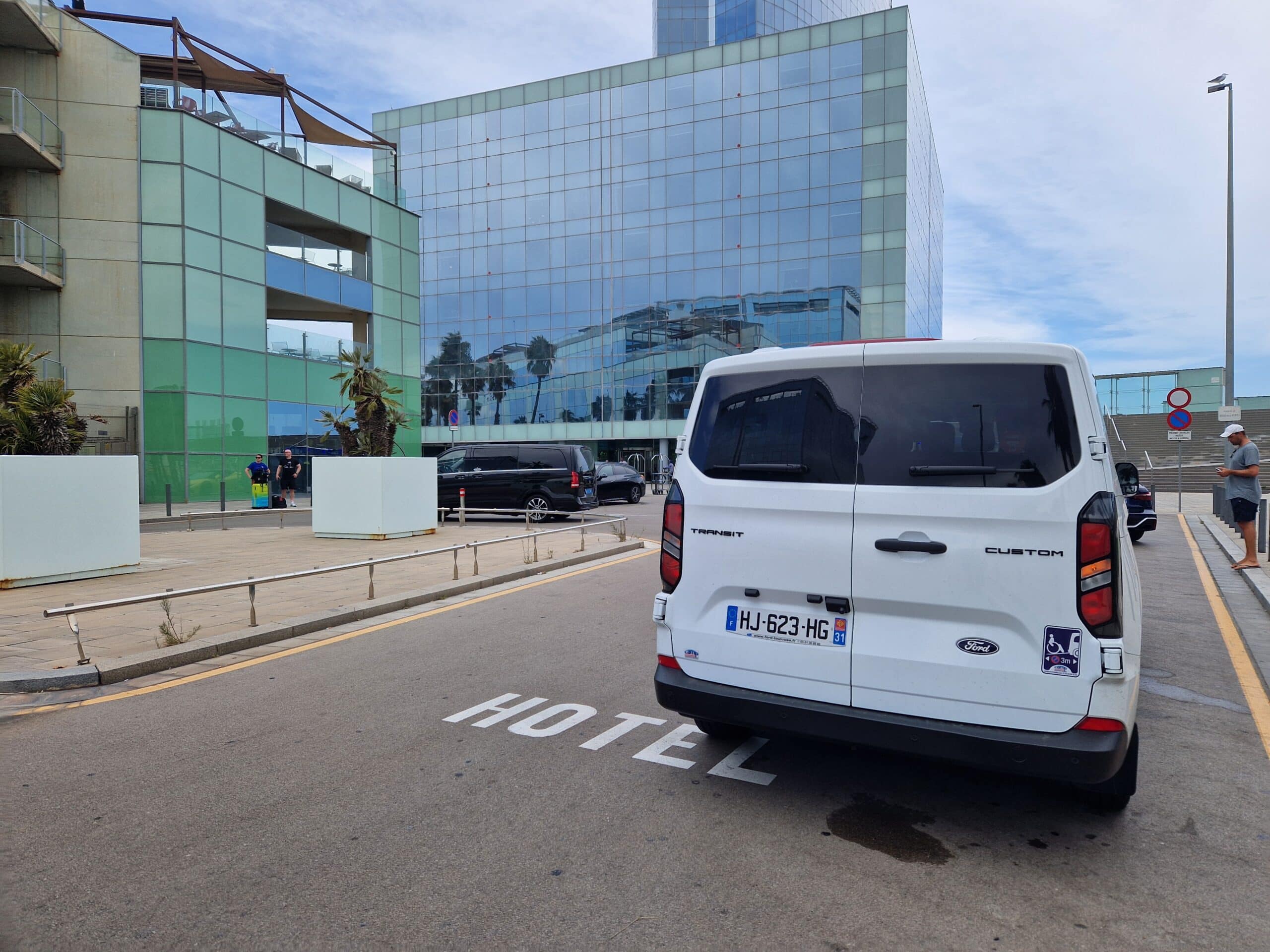 Van Ford Transit devant un hôtel moderne – Transport Van Ford Transit HJ-623-HG stationné sur marquage HOTEL devant un immeuble moderne en verre.